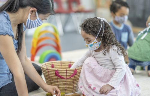 Multi ethnic group of children playing on the carpet at a daycare with a facilitator, all are wearing masks to protect from the transfer of germs after reopening.